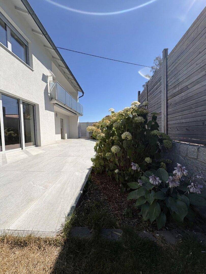 Seitlicher Gartenbereich mit gepflasterter Fläche, Blumenbeeten und Blick auf den Balkon.