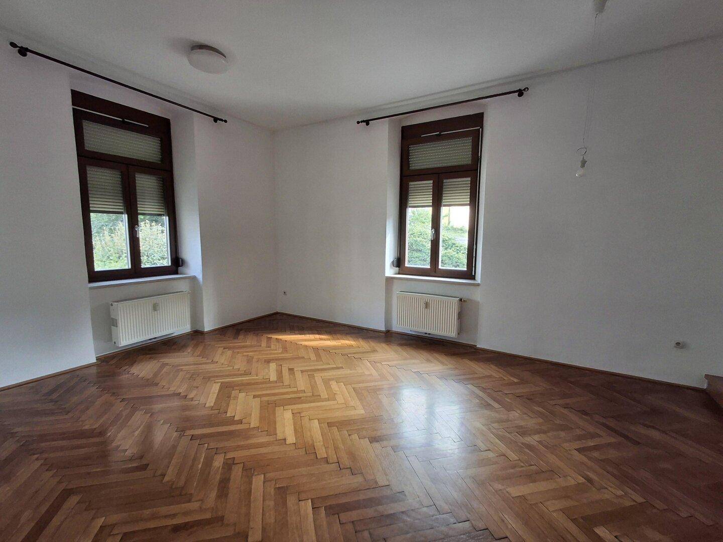 Leeres Zimmer mit Fischgrätparkett, zwei Fenstern und Heizkörpern, bereit zur Einrichtung.