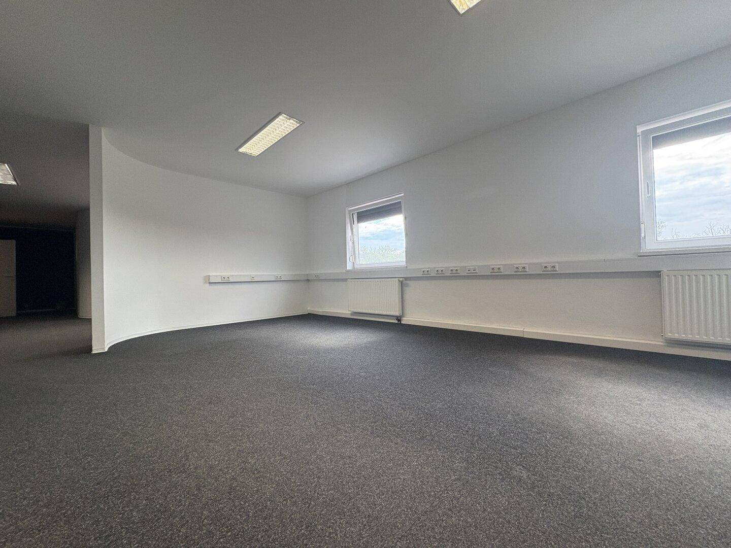 Helles Büro mit zwei Fenstern, Heizkörpern und Kabelkanälen entlang der Wände.
