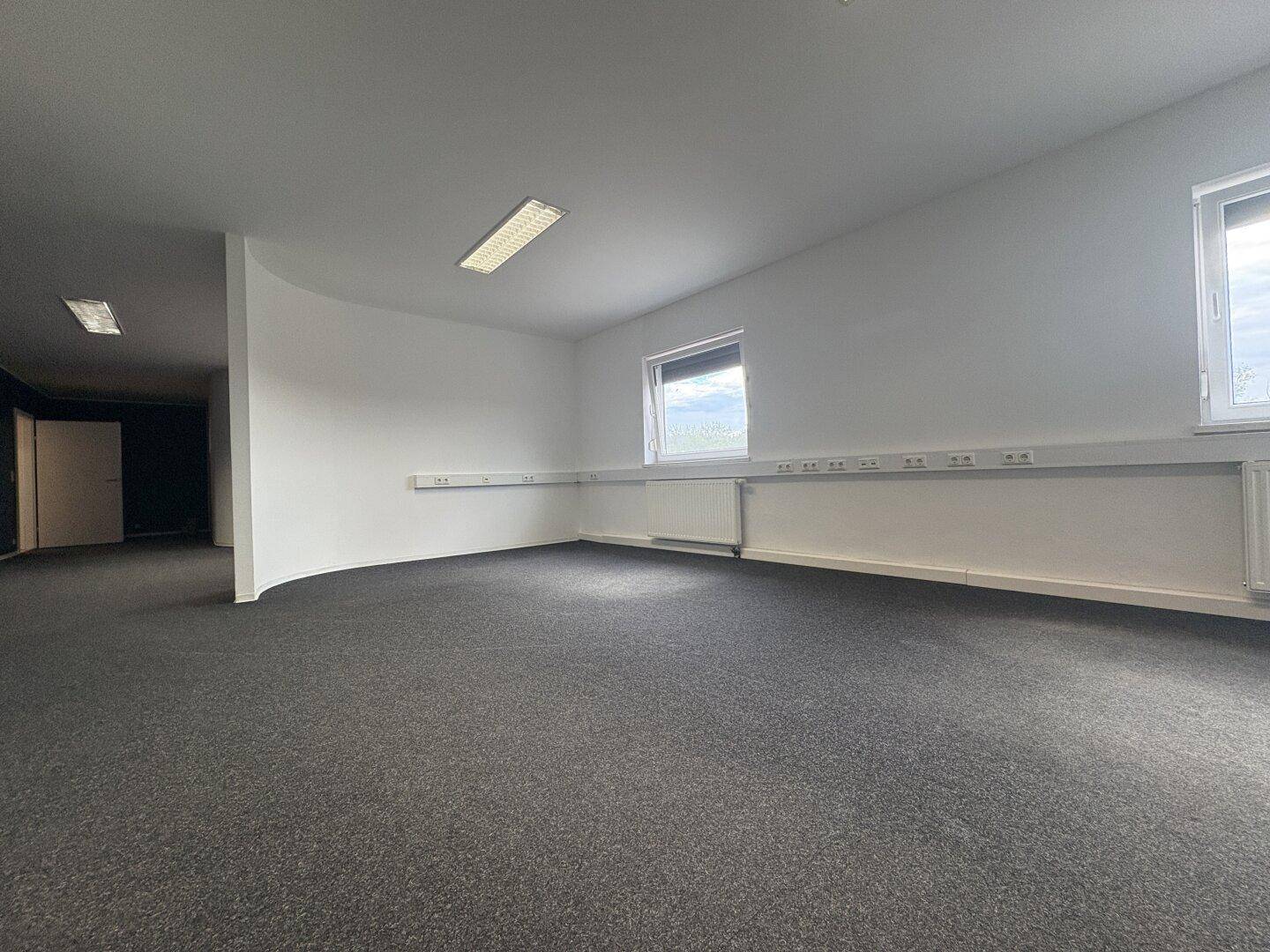 Bürofläche mit grauem Teppichboden, weißen Wänden und sichtbaren Kabelkanälen unter den Fenstern.