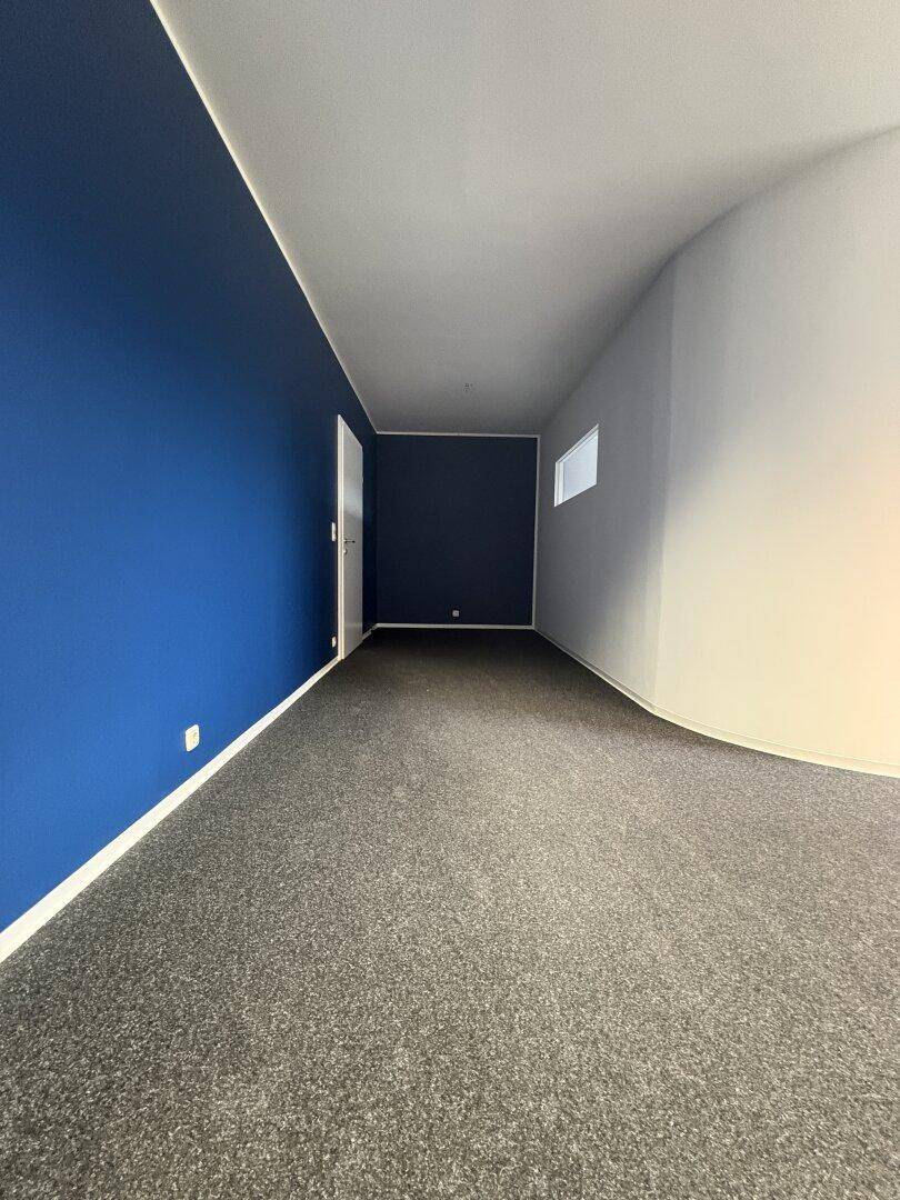 Langes Büro mit dunkelblauer Wand, grauem Teppichboden und einem kleinen Fenster.