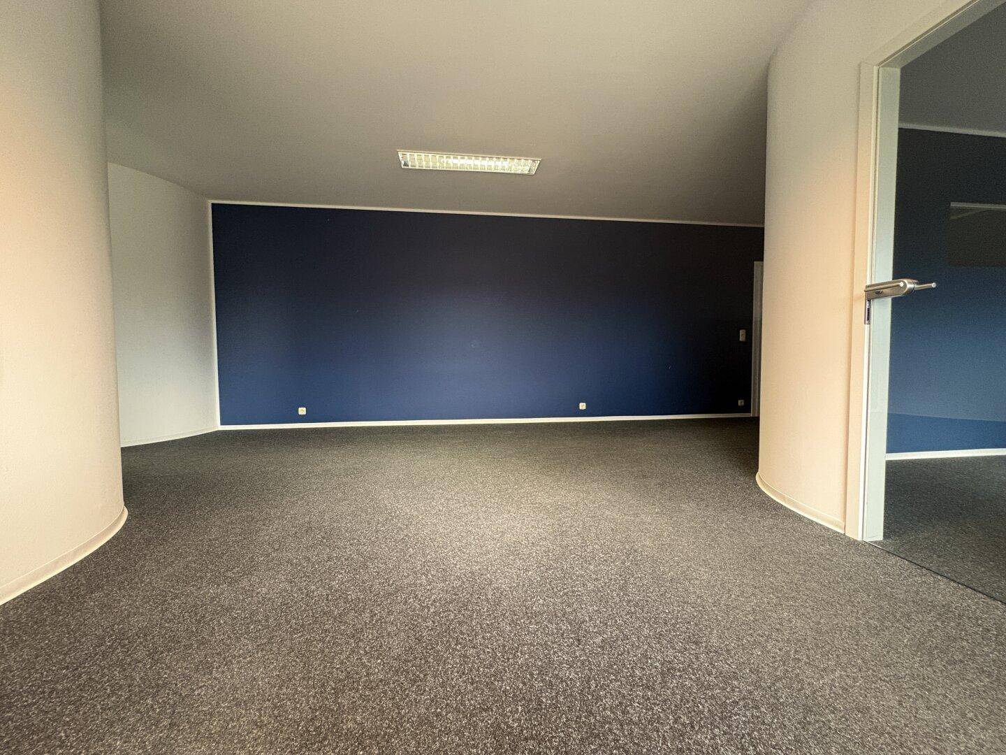 Großes Büro mit dunkelblauer Wand, grauem Teppichboden und einer Glastür zu einem Nebenraum.
