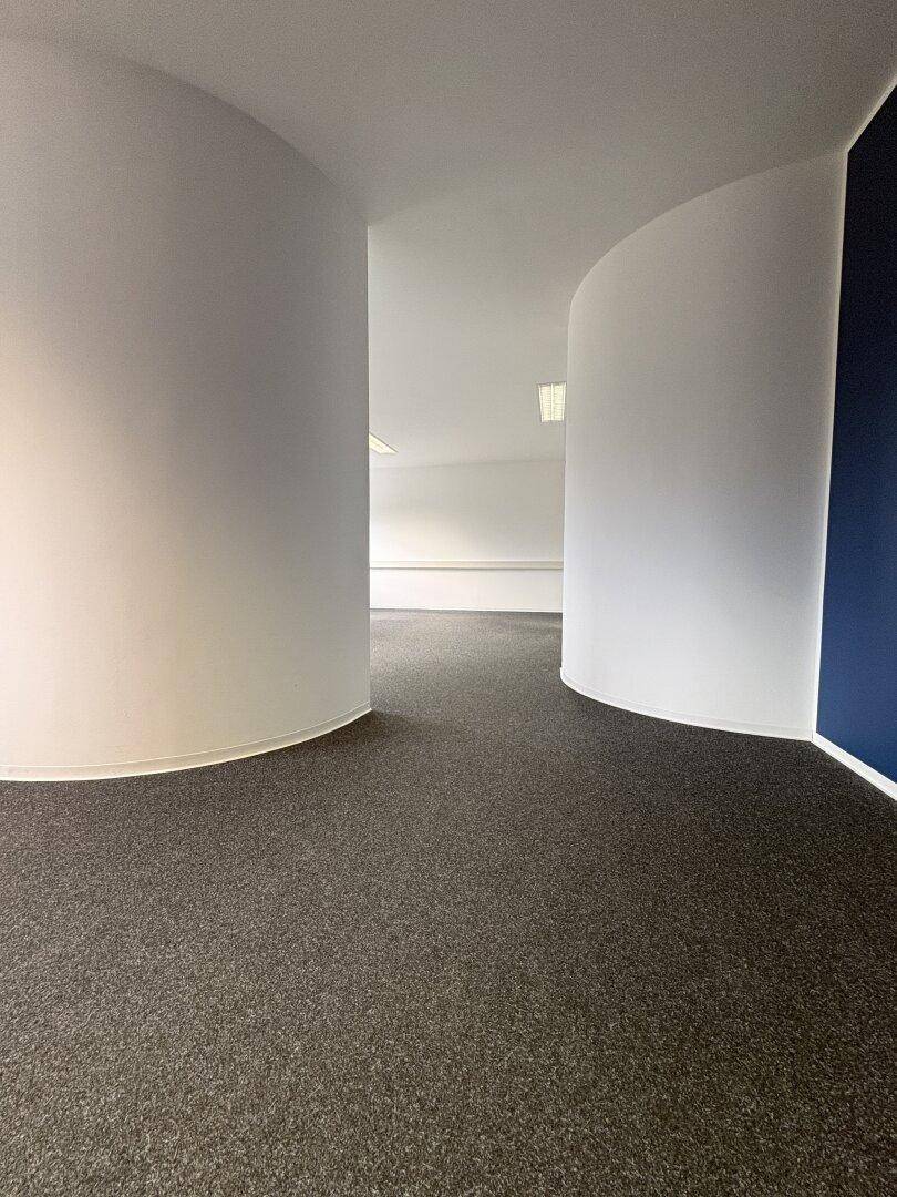 Flur oder offener Bürobereich mit geschwungenen Wänden und grauem Teppichboden.