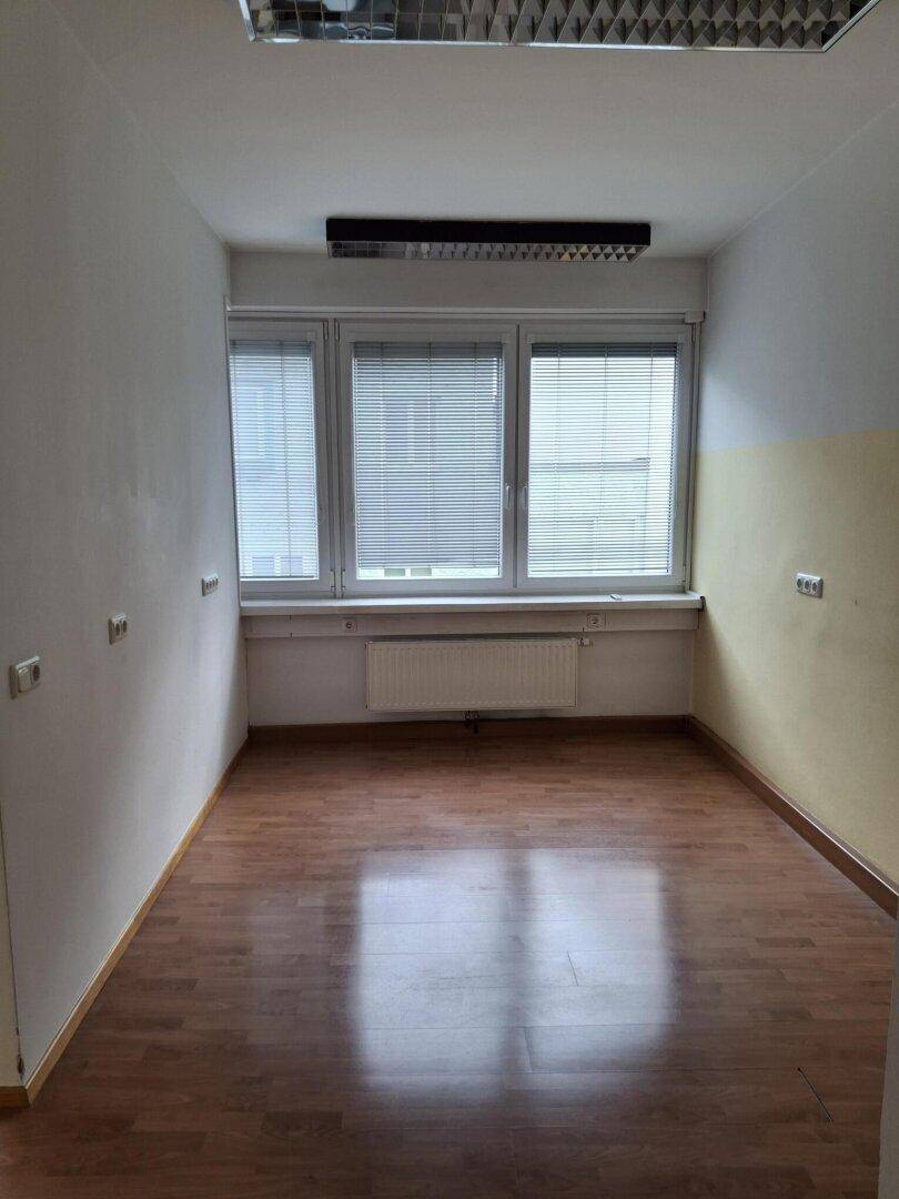 Heller Büroraum mit Laminatboden, drei Fenstern mit Jalousien und einfacher Deckenbeleuchtung.