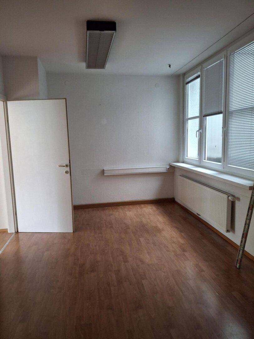 Heller Büroraum mit Laminatboden, großen Fenstern und einer Wandablage, ideal für Arbeitszwecke.