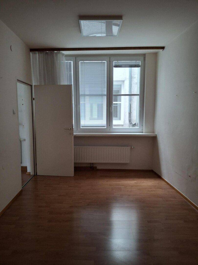Leerer Raum mit Laminatboden, zwei Fenstern mit Jalousien und einem einfachen Deckenlicht.