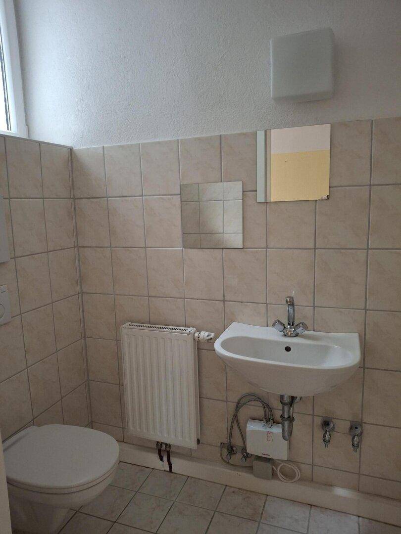 Badezimmer mit beigefarbenen Fliesen, Toilette, Waschbecken und einem Fenster mit Heizkörper.