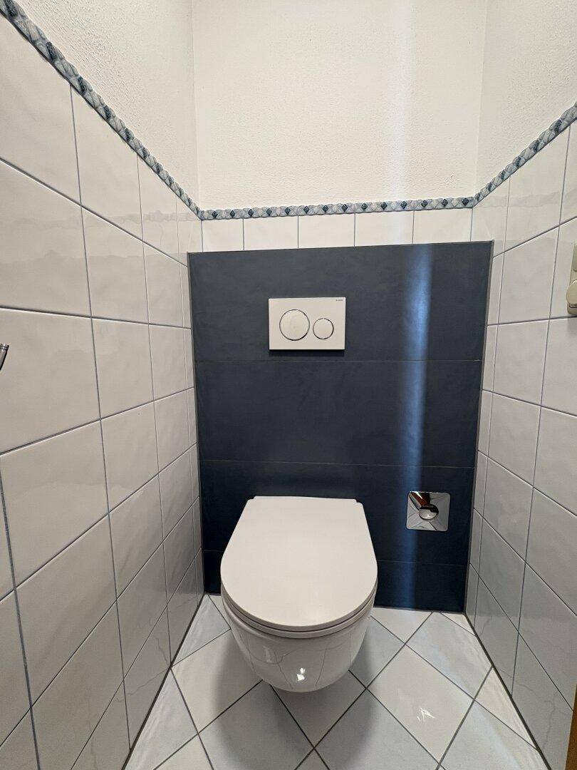 Separates WC mit Wand-WC und modernen Fliesen in Weiß und Dunkelgrau.