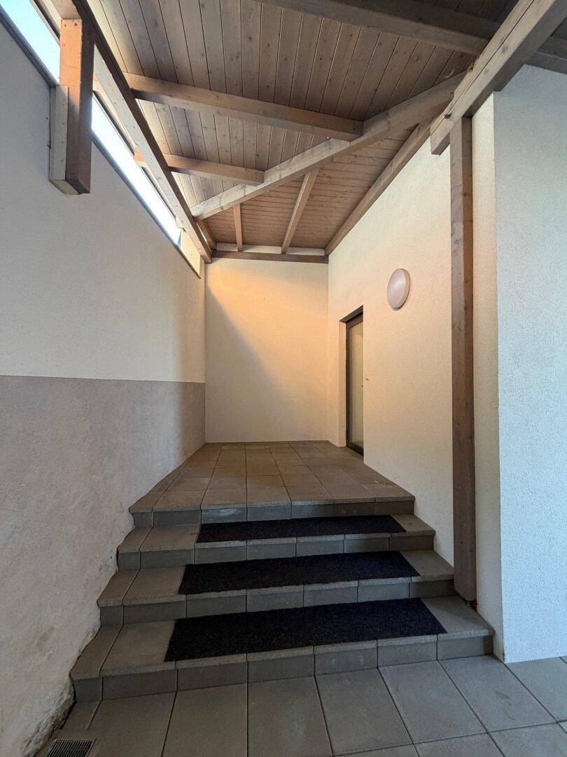 Überdachter Eingangsbereich mit Treppe, Fliesenboden und einer Holzdeckenkonstruktion.