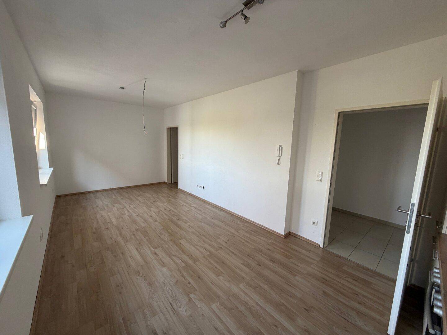 Geräumiges, leeres Wohnzimmer mit hellem Laminatboden und zwei Fenstern für viel Tageslicht.