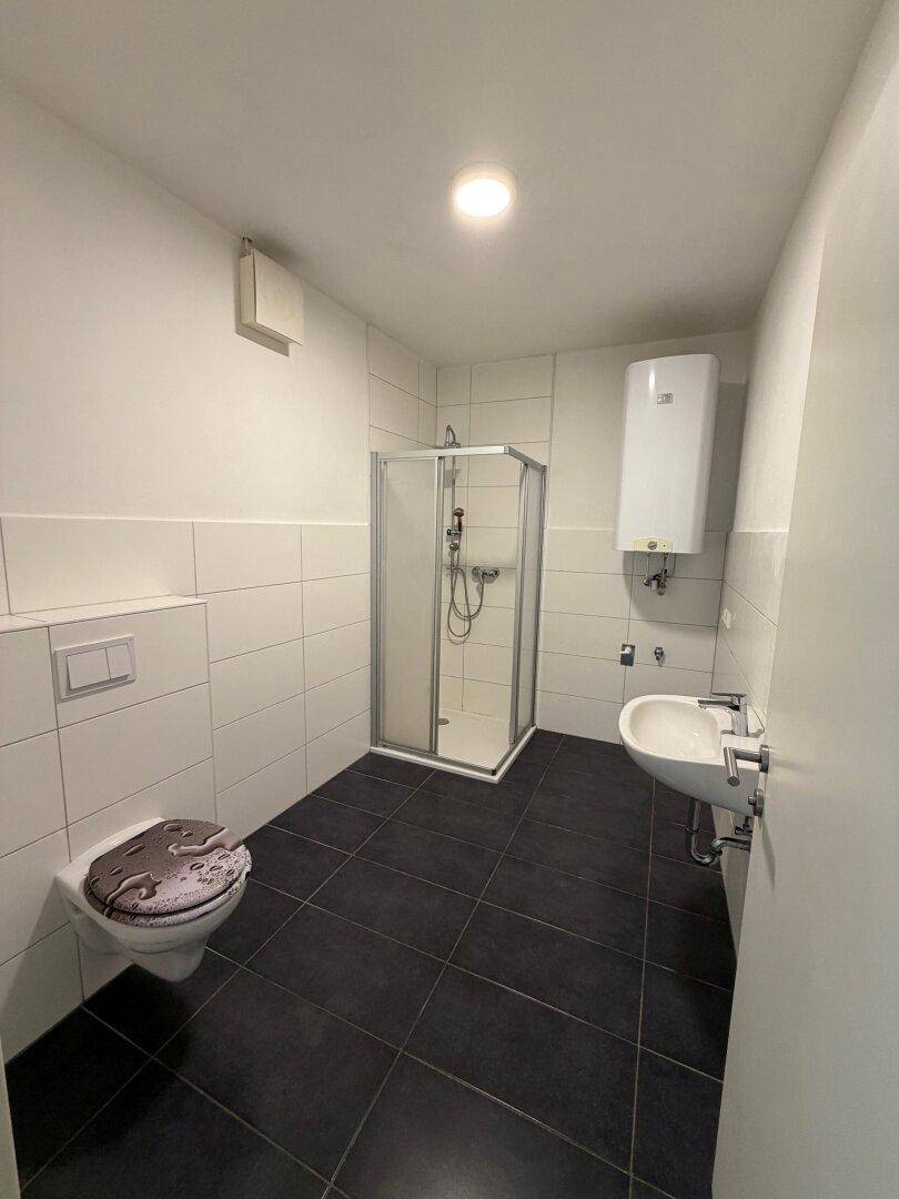 Badezimmer mit dunklen Fliesen, weiß gefliesten Wänden, Duschkabine, WC und Waschbecken.
