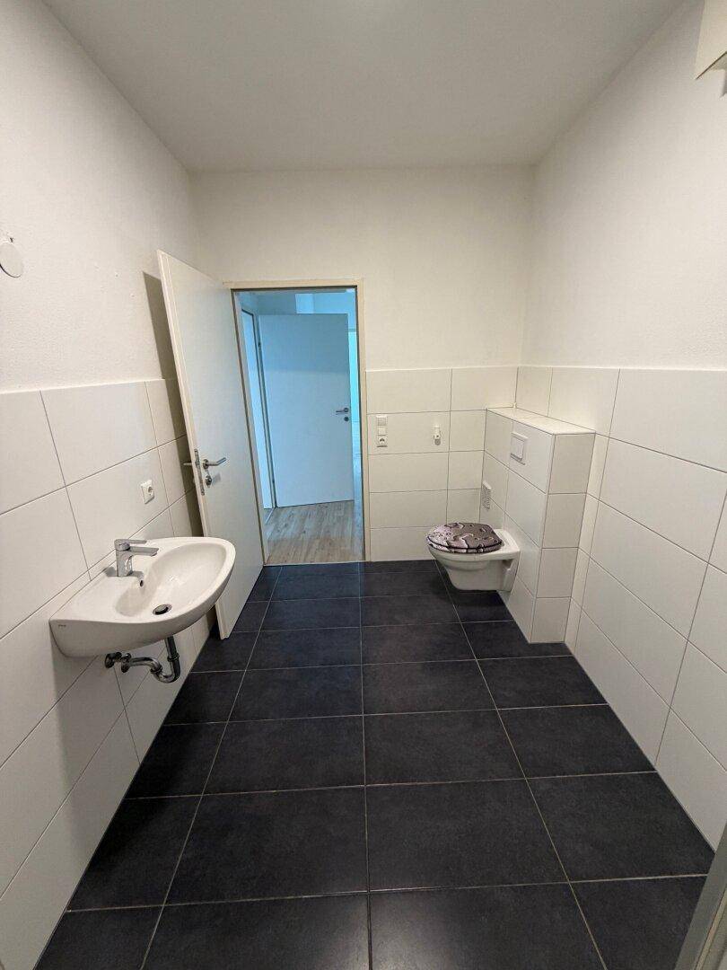 Helles Badezimmer mit dunklem Fliesenboden, weiß gefliesten Wänden, WC und Waschbecken.