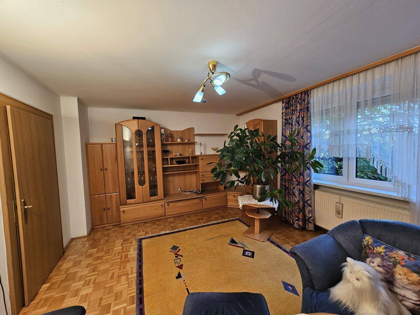 Wohnzimmer mit Parkettboden, Holzmöbeln und großem Fenster mit Vorhängen.