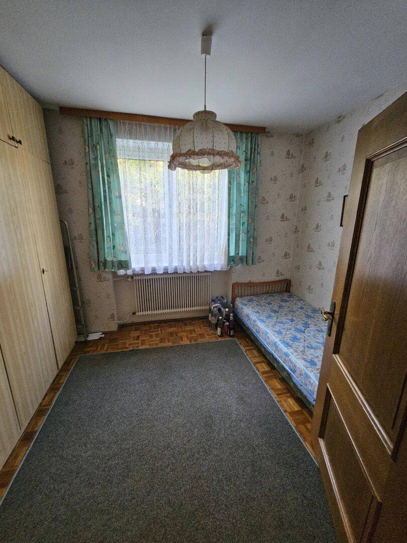 Zimmer mit Teppichboden, Einzelbett und Fenster mit Vorhängen.