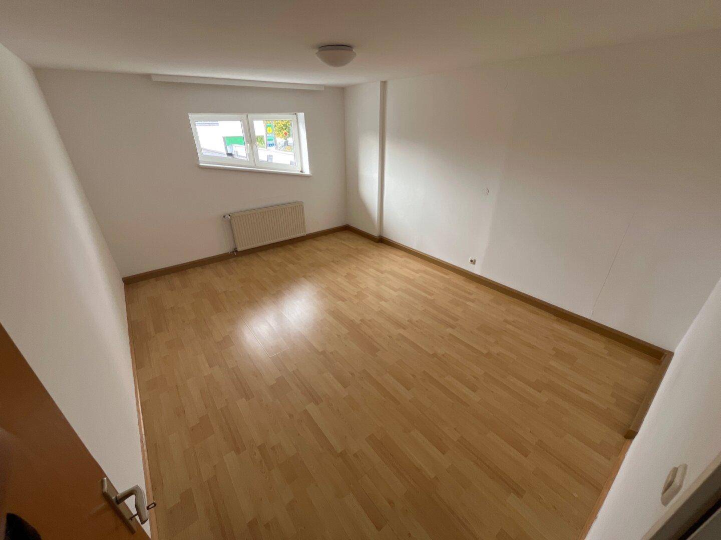 Freundliches Zimmer mit hellem Holzparkettboden, weißen Wänden und einem Fenster, das den Raum erhellt.