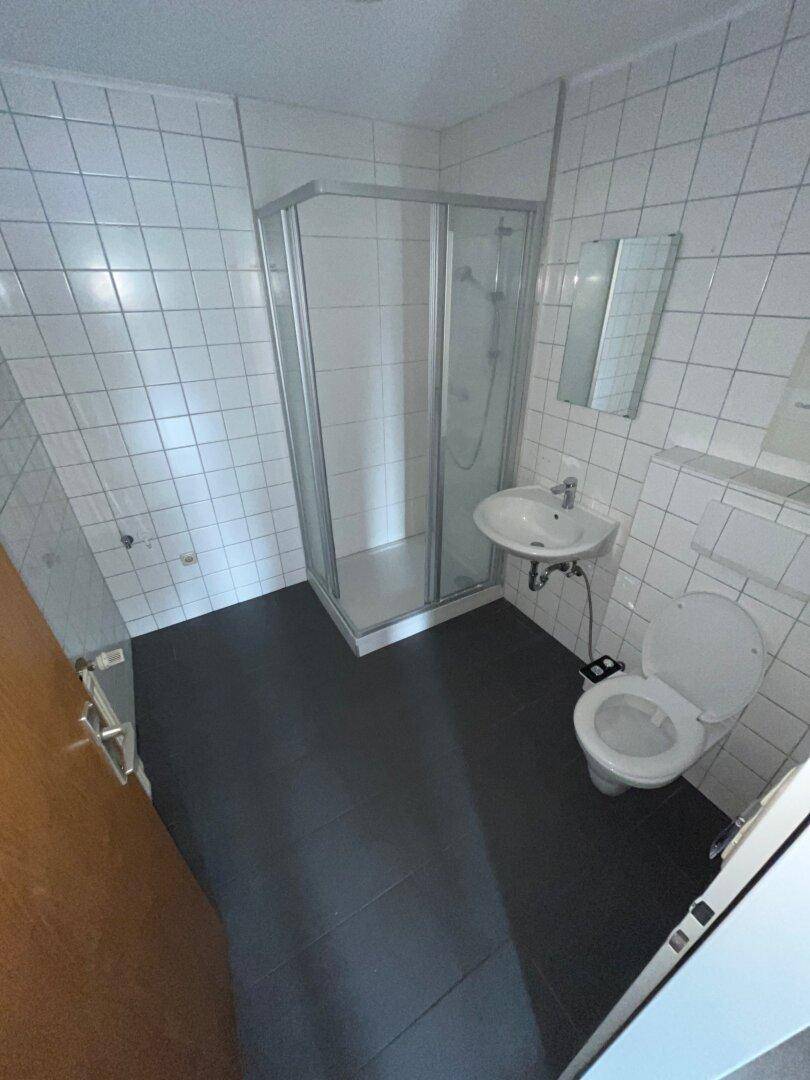 Funktionales Badezimmer mit weißen Wandfliesen, dunklem Fliesenboden, Duschkabine, Waschbecken und Toilette.