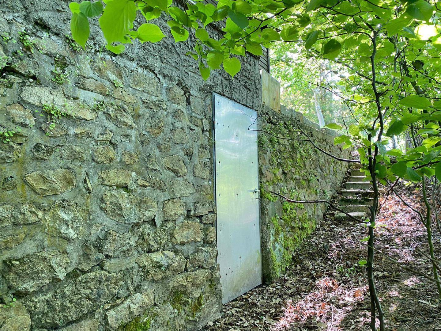 Eine robuste Steinmauer mit einer Metalltür, umgeben von üppigem Grün und einem Treppenaufgang.