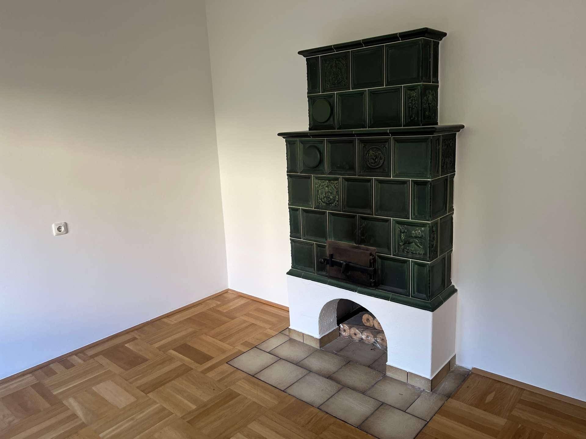Detailansicht des traditionellen grünen Kachelofens mit Holzlagerfach in einem hellen Raum.