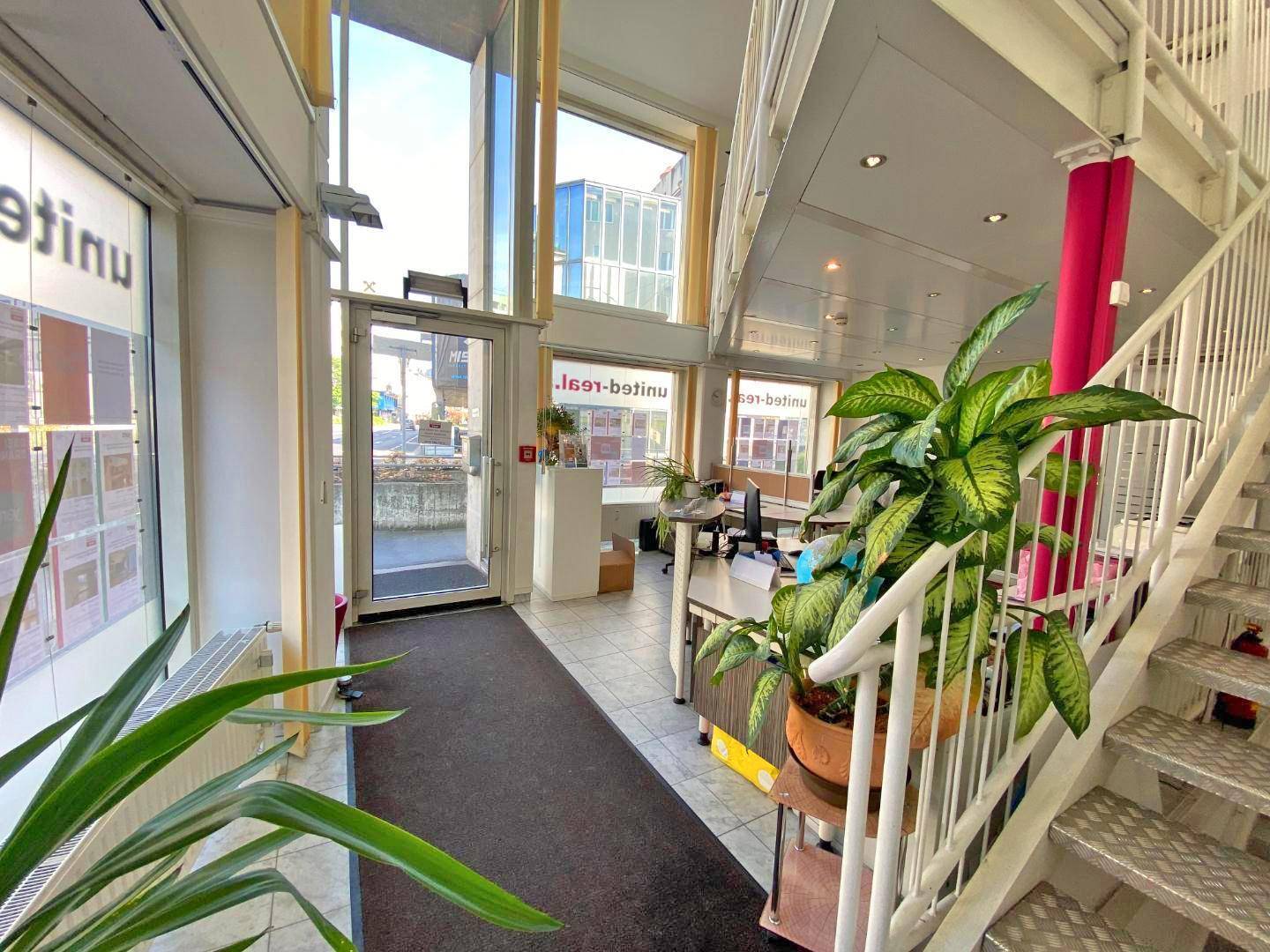 Eingangsbereich eines Büros mit Treppe, großen Fenstern und einem Empfangstresen, der viel Tageslicht erhält.