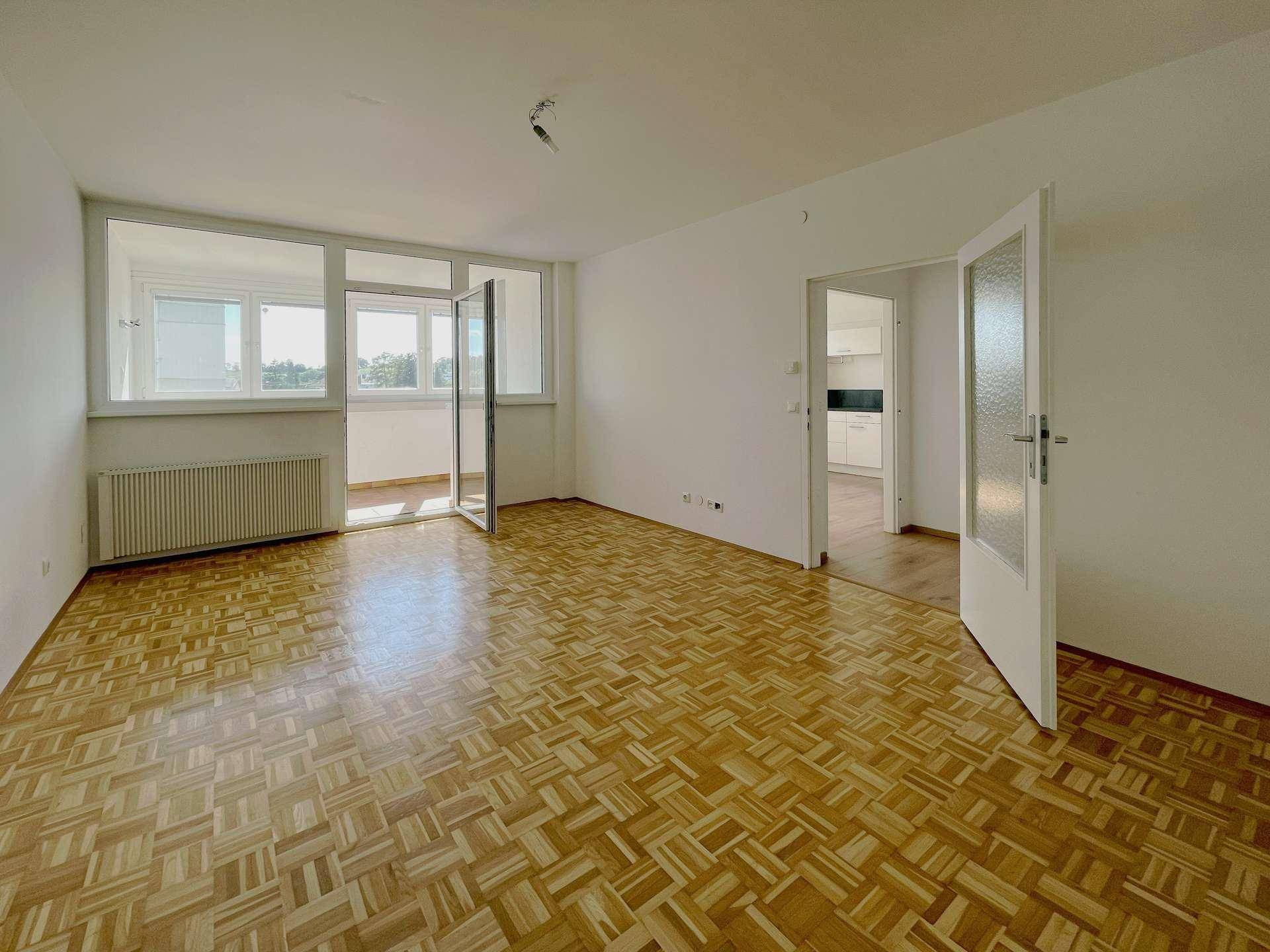 Helles Wohnzimmer mit Parkettboden, weißen Wänden und Zugang zu einem Balkon.