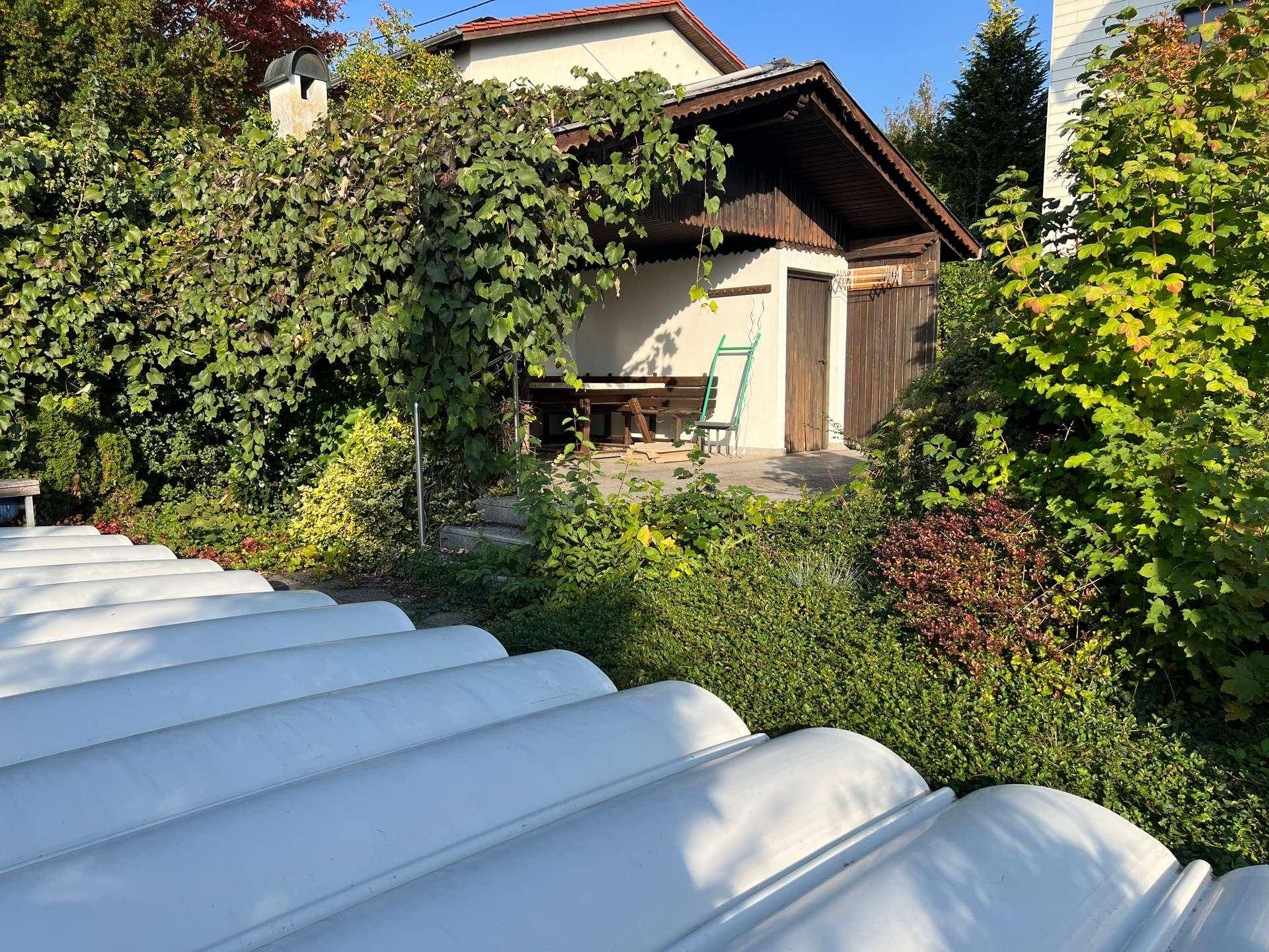 Überdachter Außenbereich mit Sitzgelegenheiten und Grillplatz, umgeben von Grün, mit Poolabdeckung im Vordergrund.