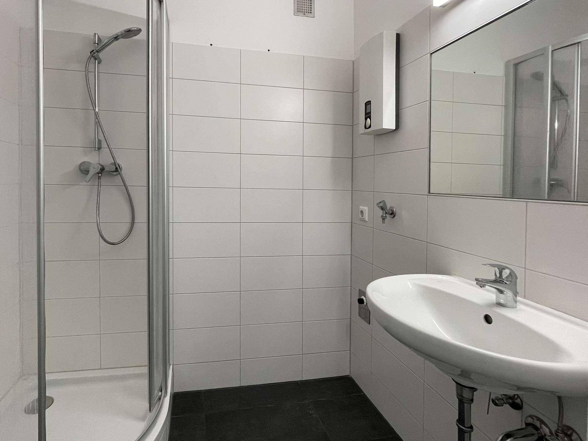 Helles Badezimmer mit Dusche, Waschbecken, Spiegel und weißen Fliesen.