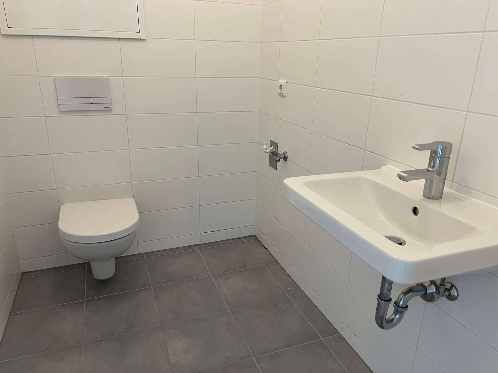 Modernes Gäste-WC mit weißen Wandfliesen, grauem Boden und wandhängendem WC sowie einem Waschbecken.