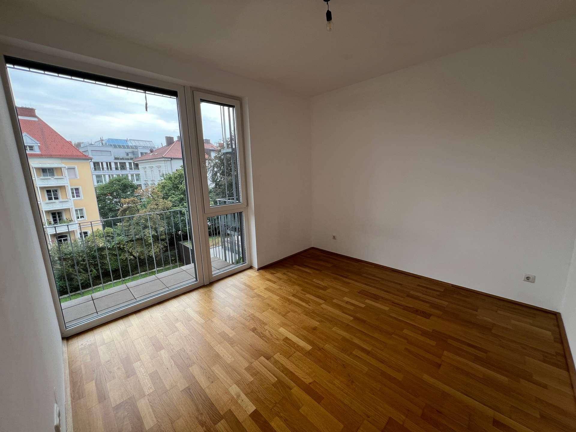 Helles Zimmer mit Holzboden und großen Fenstern, die Zugang zu einem französischen Balkon bieten und viel Tageslicht hereinlassen.