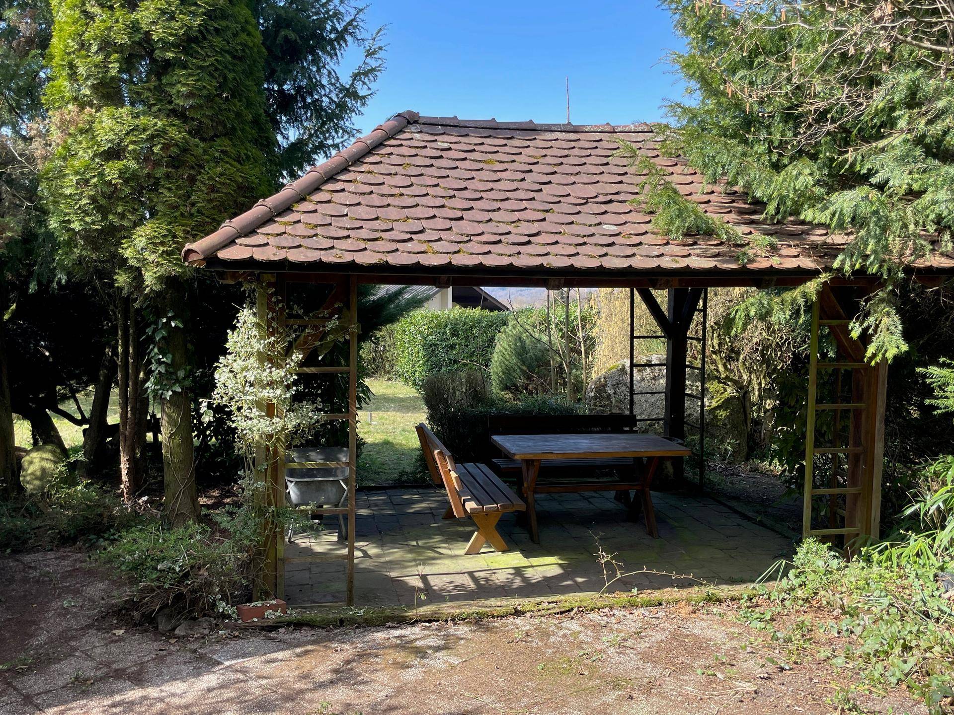 Gemütlicher Gartenpavillon mit Holztisch und Bänken, umgeben von üppigem Grün.
