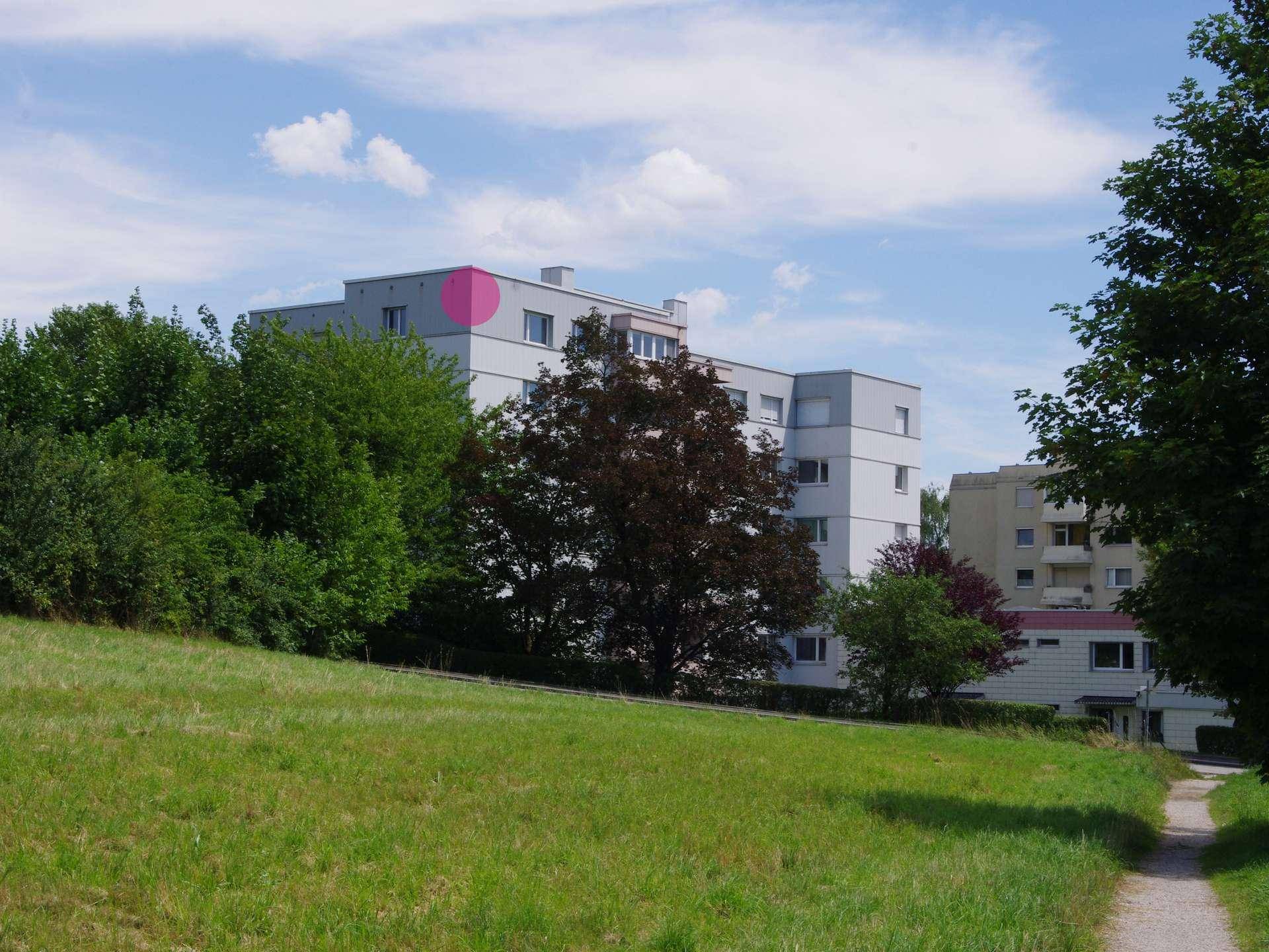Mehrfamilienhaus in einer grünen Umgebung, gesehen von einem grasbewachsenen Hügel.
