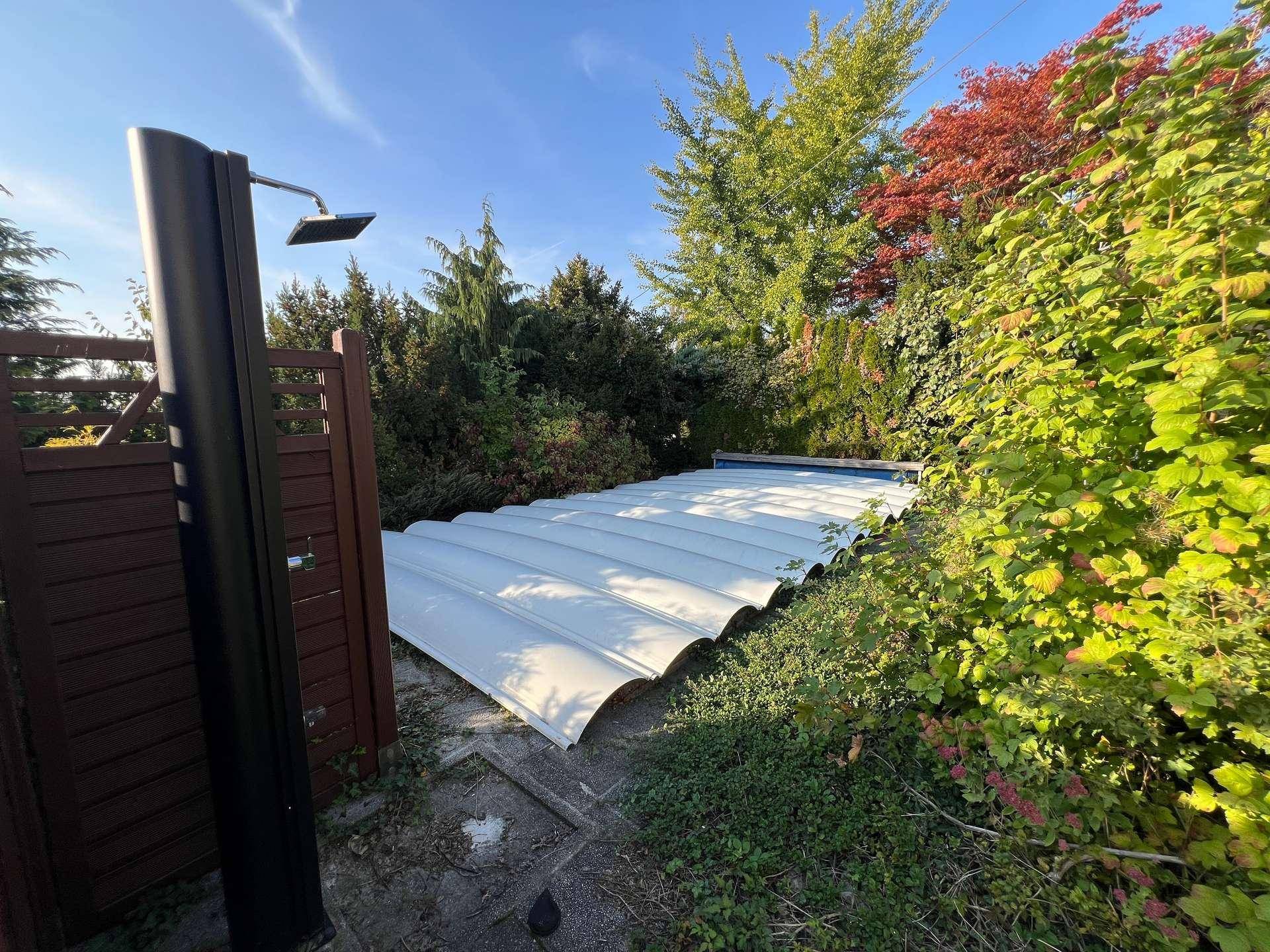 Außendusche neben einer abgedeckten Poolanlage, umgeben von üppigem Grün im Garten.