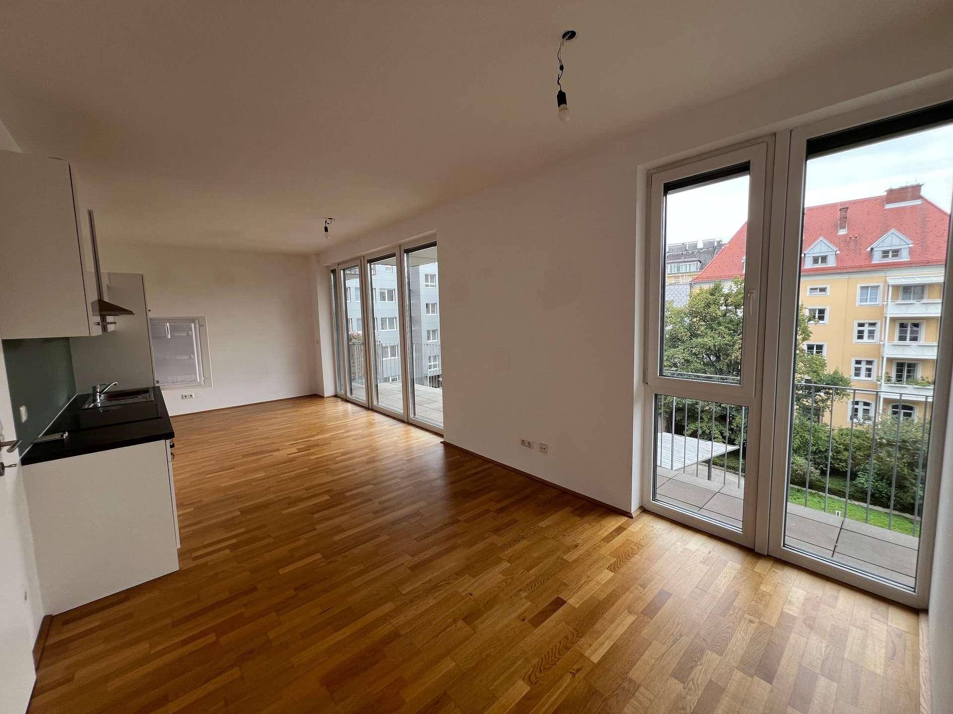 Geräumiger offener Wohnbereich mit Holzboden, integrierter Küche und großen Fenstern, die viel Licht hereinlassen.