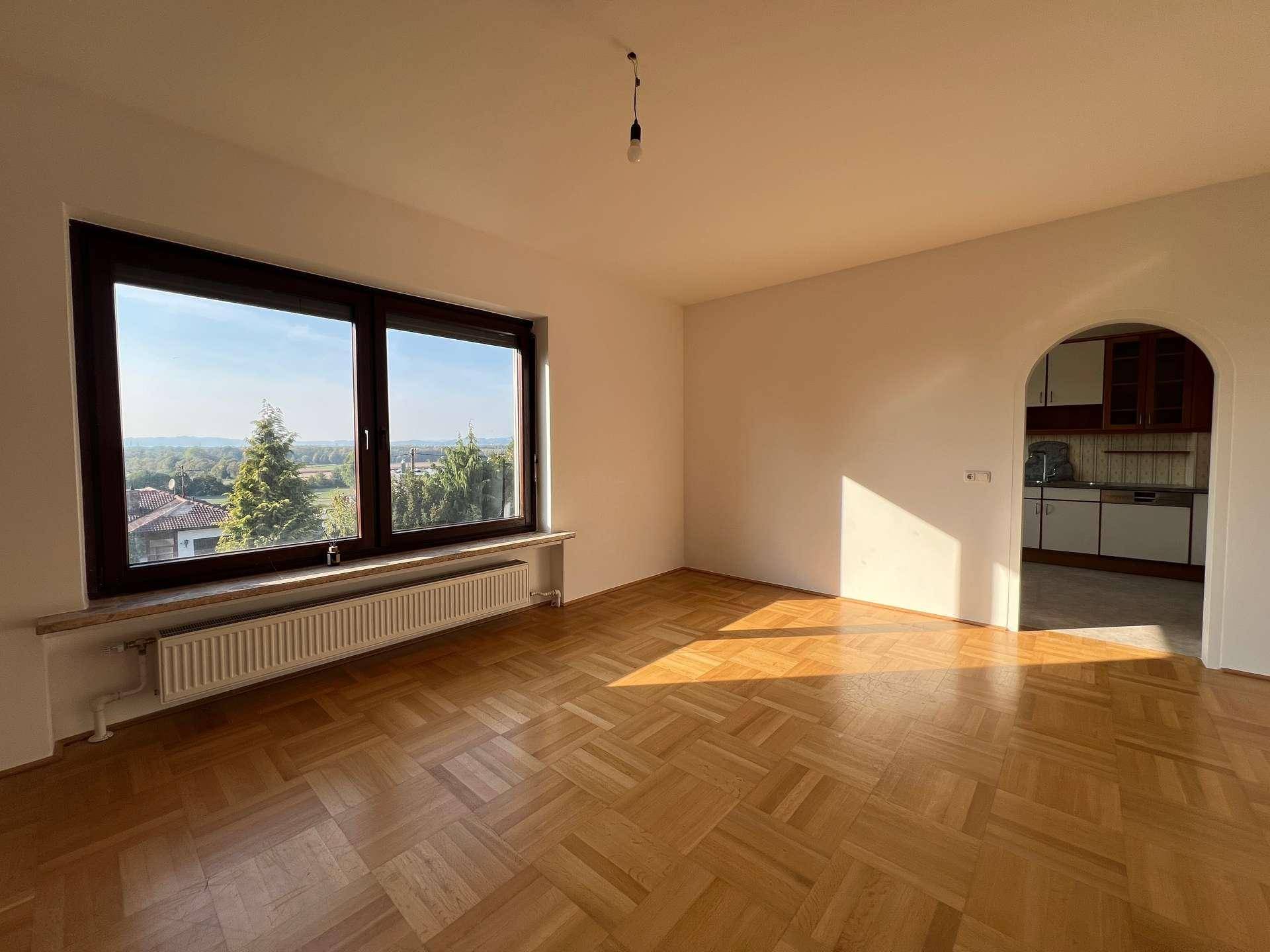 Lichtdurchflutetes Wohnzimmer mit Parkettboden, großen Fenstern und Zugang zu einer Küche durch einen Bogen.