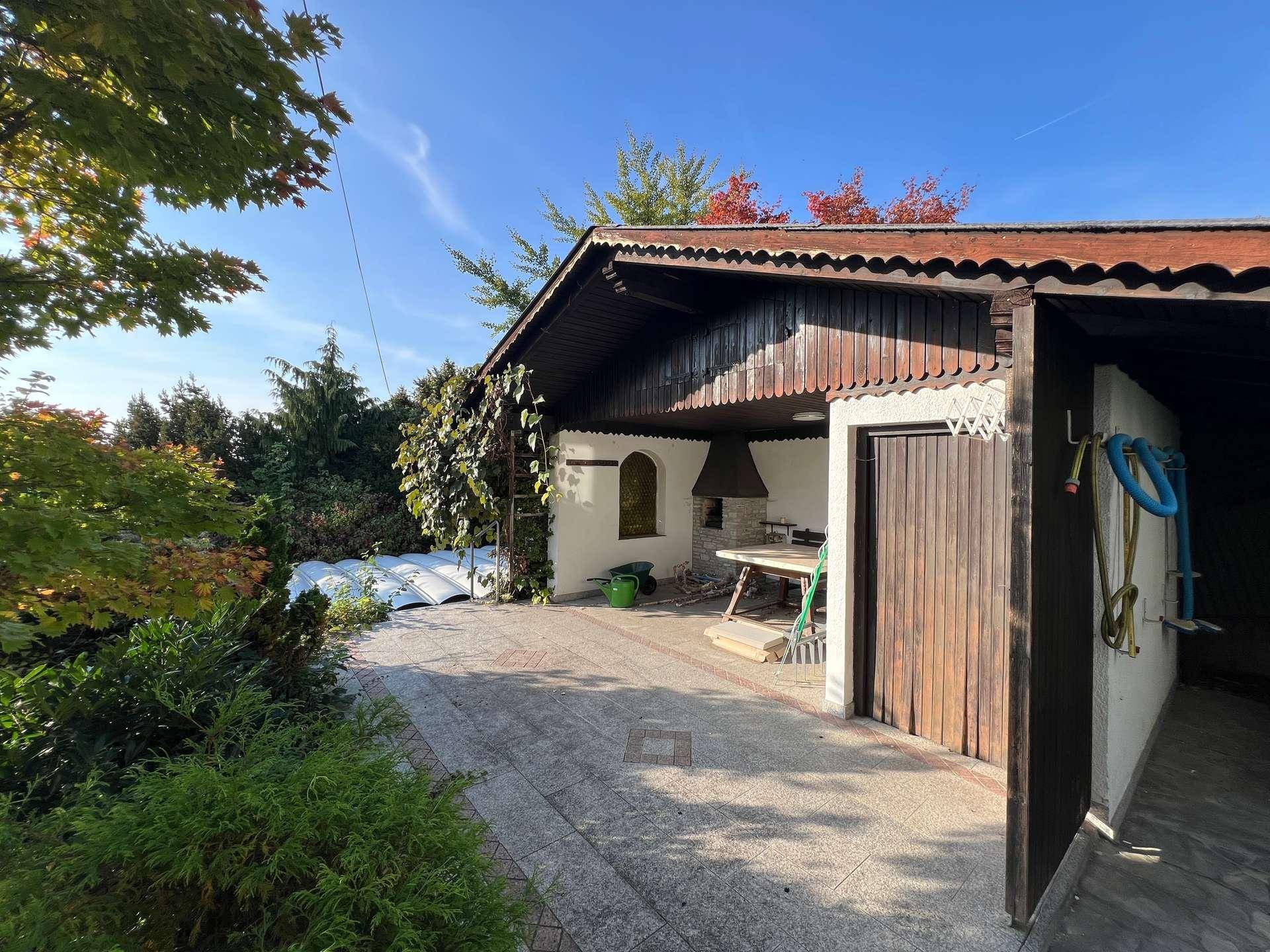 Überdachter Außenbereich mit Grillplatz, Holzelementen und üppiger Bepflanzung im Garten.