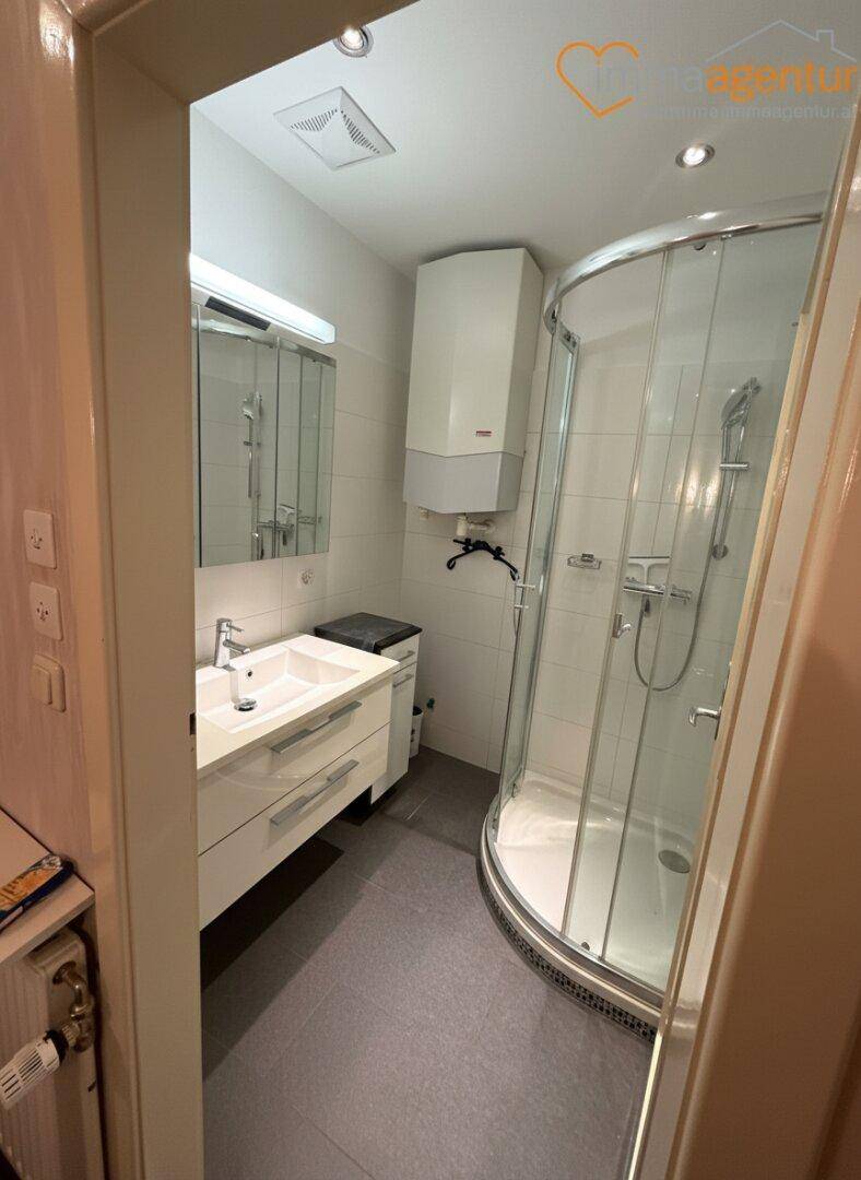 Kleines Badezimmer mit Duschkabine, Einzelwaschbecken und einem Warmwasserbereiter an der Wand.