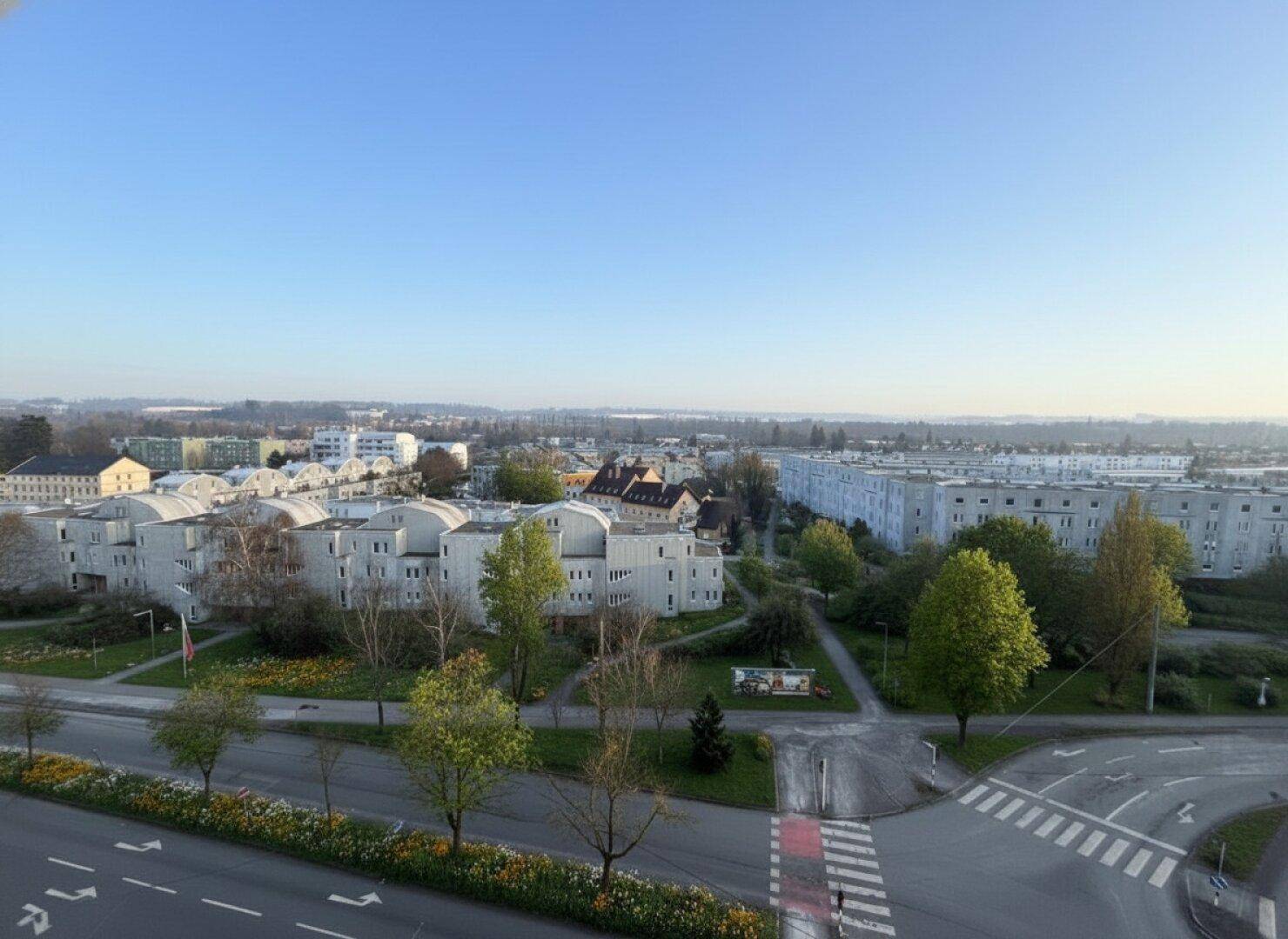 Weitläufiger Blick über eine Stadtlandschaft mit Wohngebäuden, Straßen und Grünflächen unter blauem Himmel.