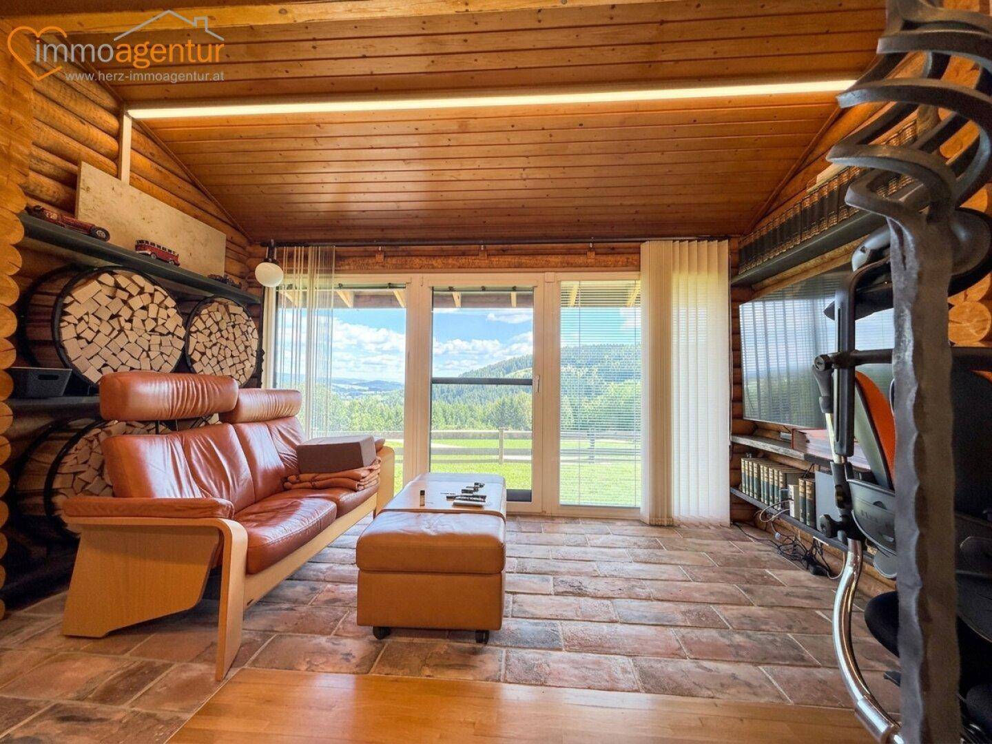Gemütliches Wohnzimmer im Blockhausstil mit Ledersofa, Kaminholzregal und weitem Blick in die Natur.