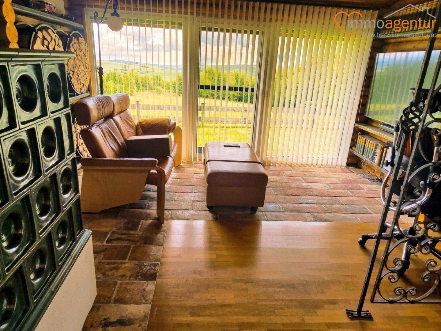 Wohnzimmer mit Kachelofen, Ledersofa und großen Fenstern, die einen weiten Blick in die Natur bieten.