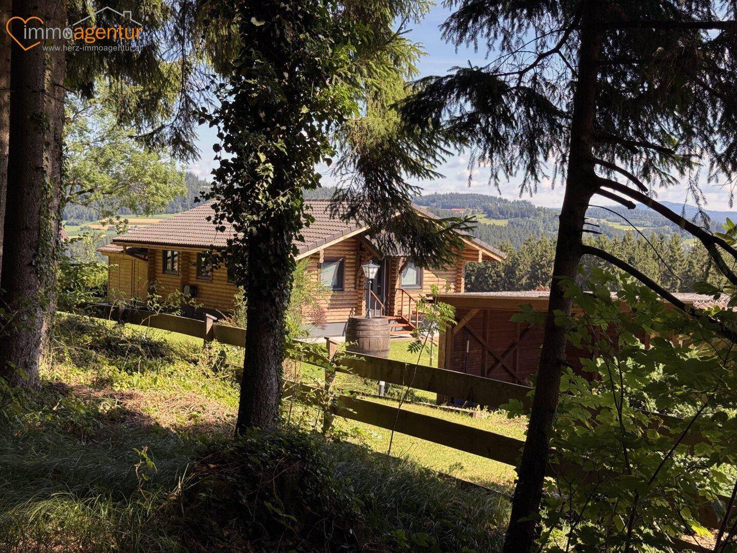 Blockhaus eingebettet in grüne Landschaft, umgeben von Bäumen und mit Blick auf die Hügel.