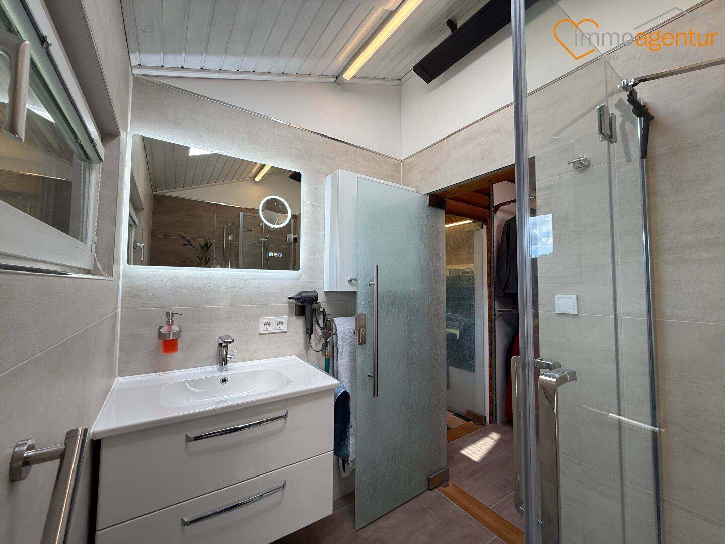 Helles Badezimmer mit modernem Waschtisch, beleuchtetem Spiegel und begehbarer Dusche.