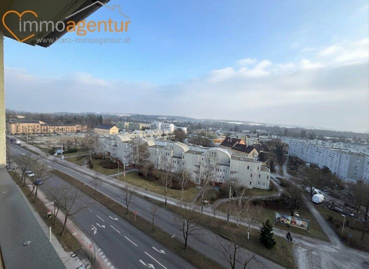 Weitläufiger Blick über eine städtische Landschaft mit Straßen und Gebäuden unter blauem Himmel.