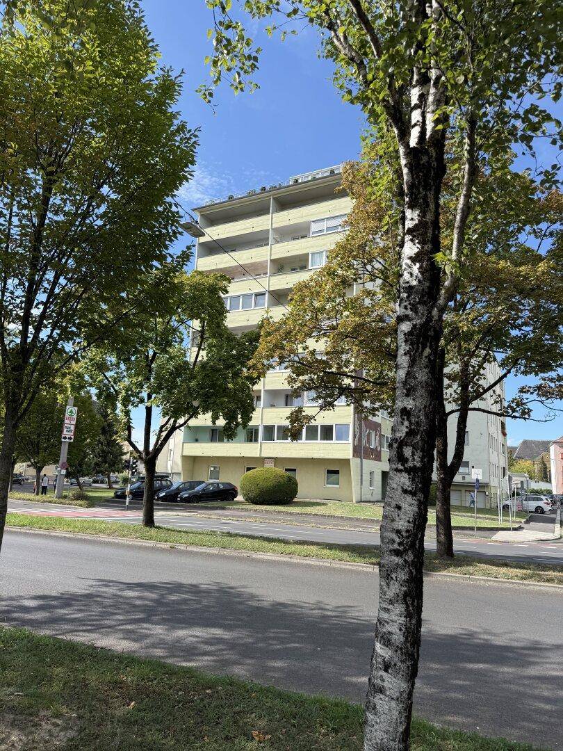 Mehrstöckiges Wohnhaus mit Balkonen, teilweise von Bäumen verdeckt, an einer Straße gelegen.