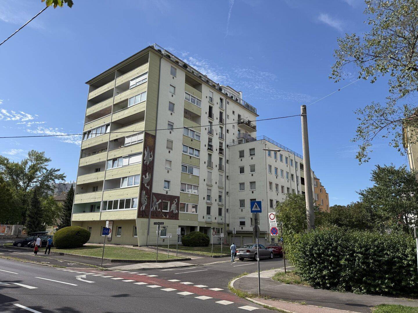 Großes Wohngebäude mit mehreren Etagen und Balkonen an einer Straßenecke mit Grünflächen.
