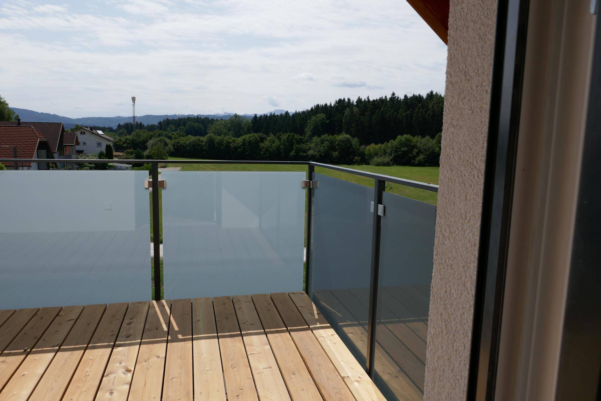 Moderner Balkon mit Holzdielenboden, elegantem Glasgeländer und einem weiten Blick in die grüne Landschaft.
