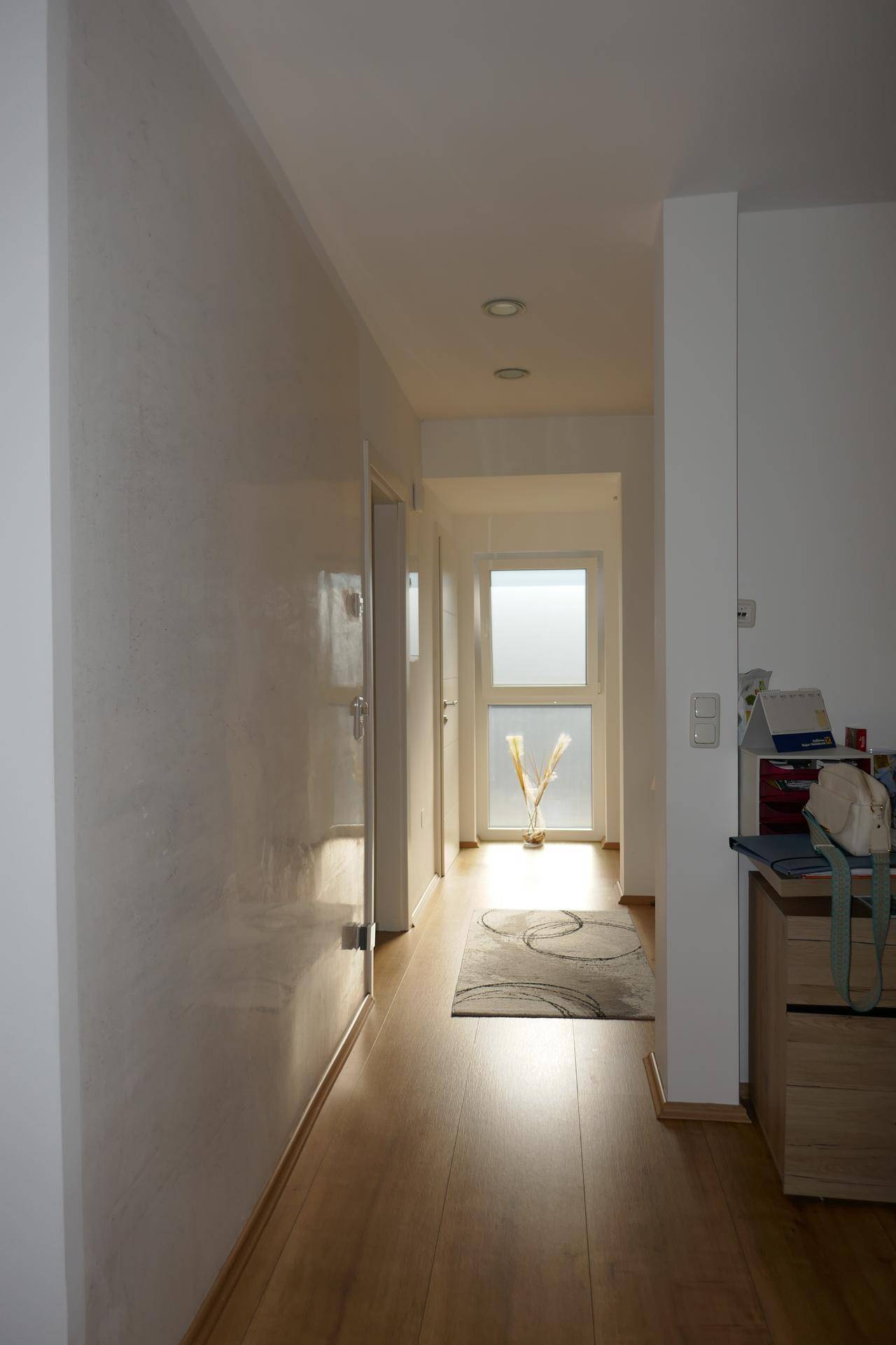 Langer, heller Flur mit Holzboden und einer modernen Tür am Ende, die Tageslicht hereinlässt.