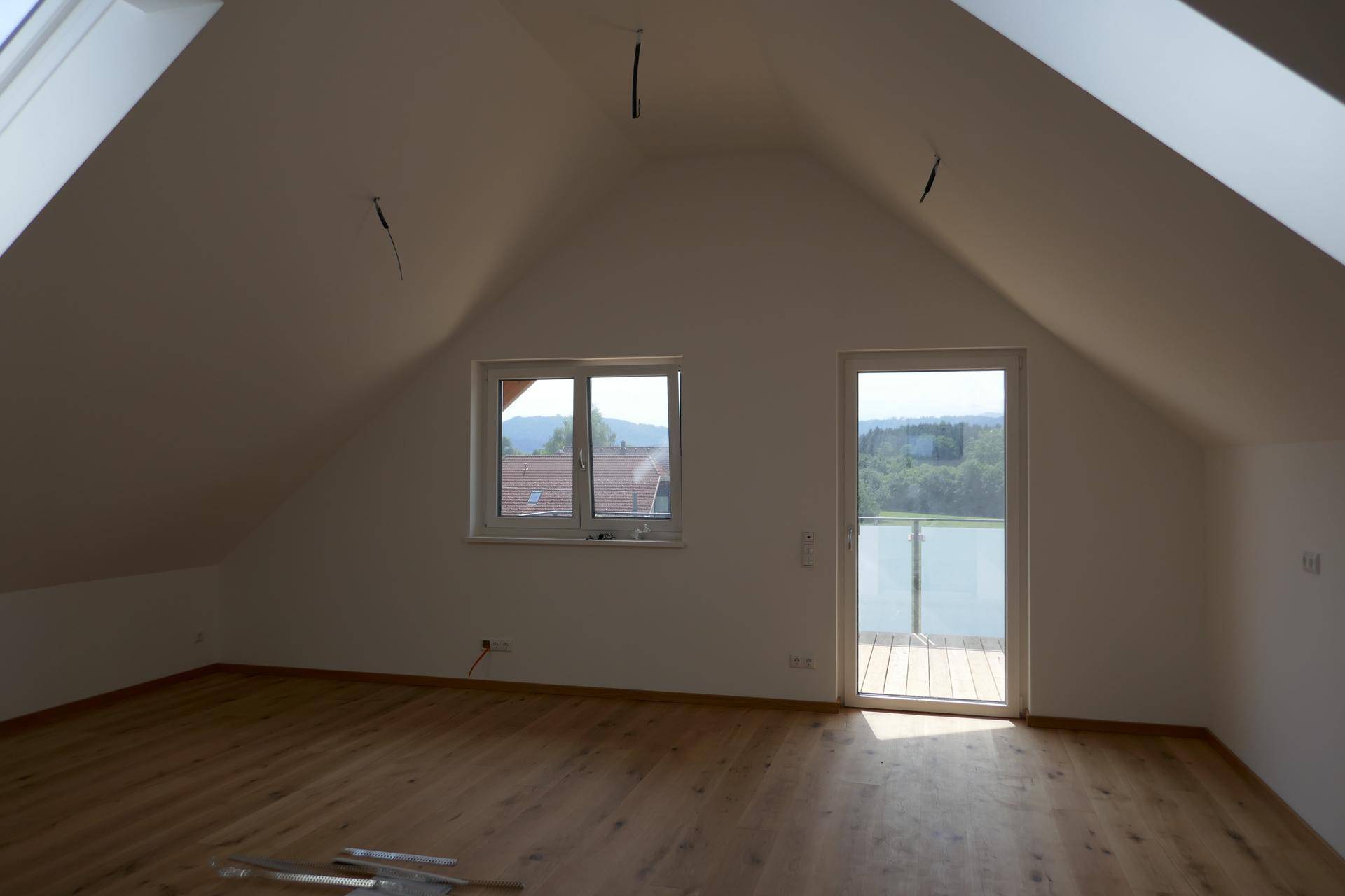 Leerer Raum im Dachgeschoss mit Holzdielenboden, schrägen Decken und Zugang zu einem Balkon mit Weitblick.
