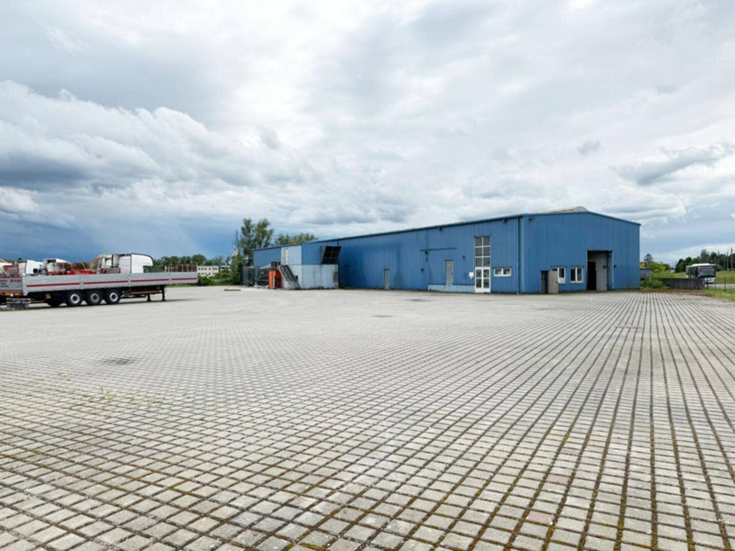 Großes blaues Industriegebäude mit weitläufiger gepflasterter Außenfläche und einem LKW-Anhänger unter bewölktem Himmel.