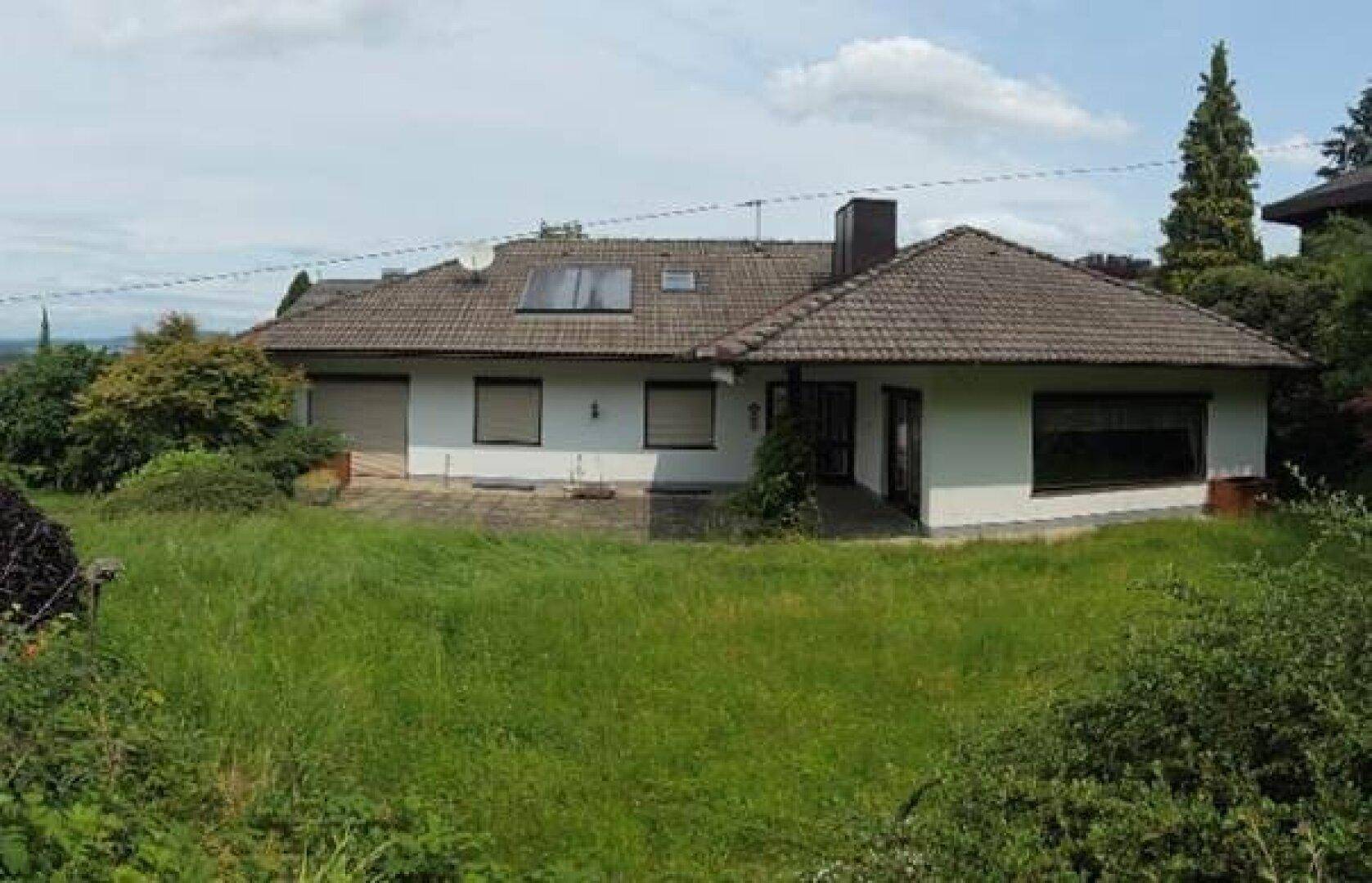 Rückseite des Hauses mit Terrasse und großem, ungemähtem Gartenbereich unter bewölktem Himmel.