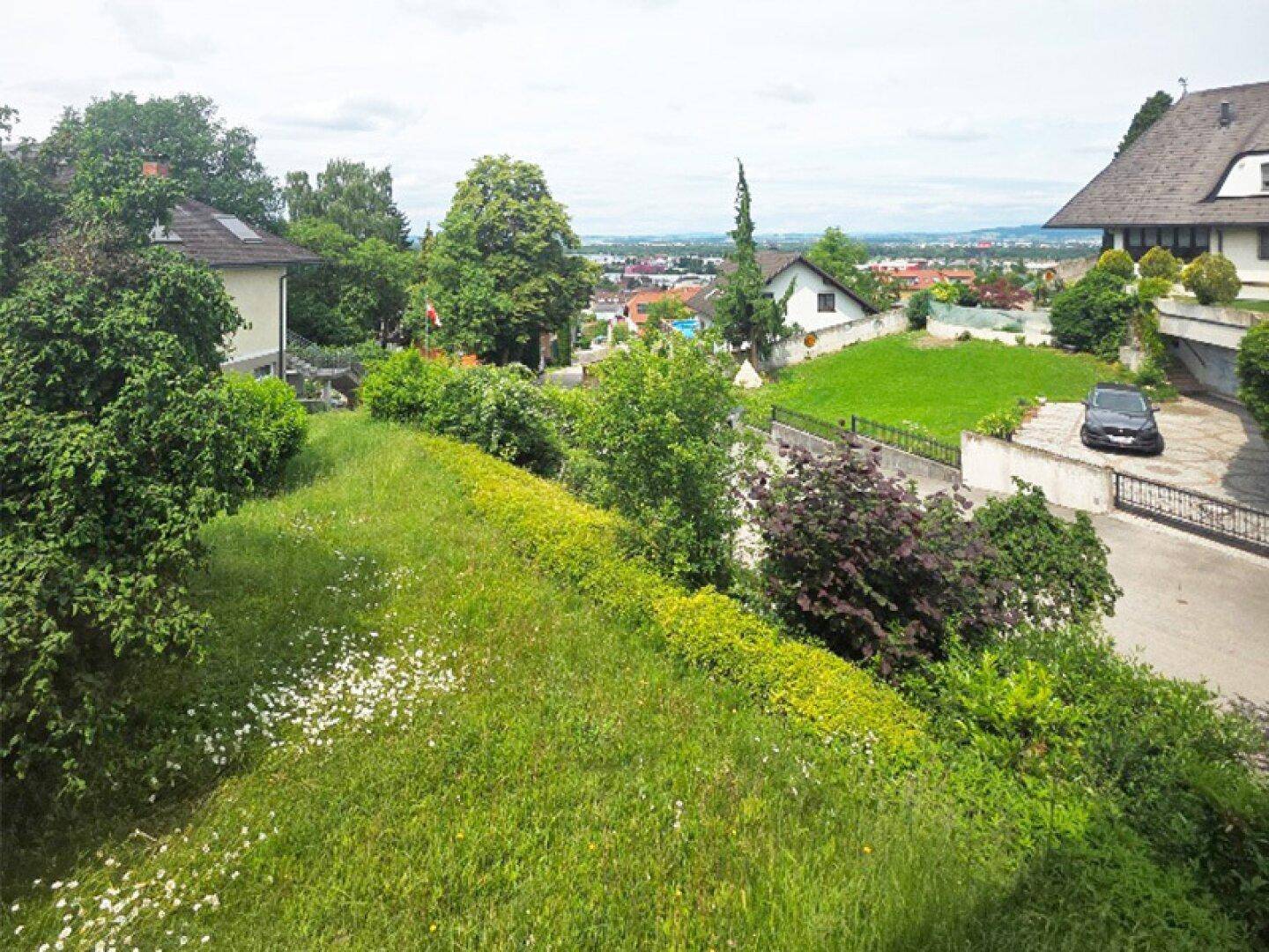 Panoramablick über eine grüne Wohngegend mit Häusern, Gärten und einer weiten Landschaft im Hintergrund.