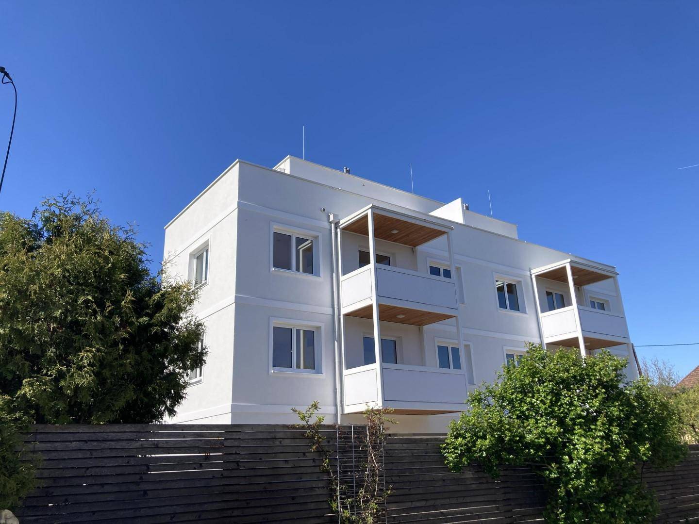 Modernes weißes Apartmentgebäude mit mehreren Balkonen unter blauem Himmel.